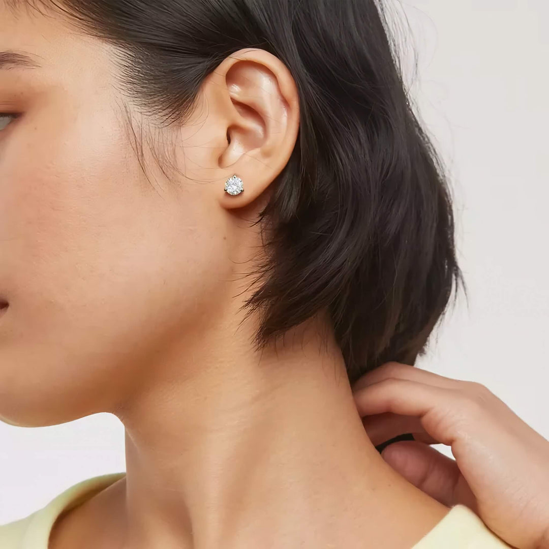 Close-up of a person wearing a diamond earring with a neutral background. Lab-grown diamond martini (3-prong) stud earrings, 2 ctw, 14K yellow gold, on ear.