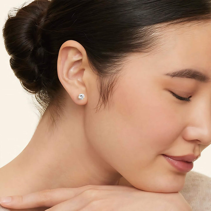 Close-up of a woman wearing a diamond earring with a neutral background. Lab-grown diamond martini (3-prong) stud earrings, 2 ctw, 14K yellow gold, on model.